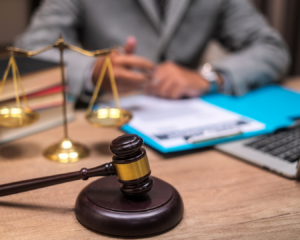 Close up shot of lawyer's desk with gavel, scales of justice, paperwork, books, and laptop. Male lawyer can be seen in background, wearing a suit and going over documents.