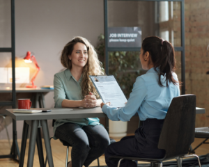 Young woman sitting across desk from female manager who is going over her application. Inside modern office.